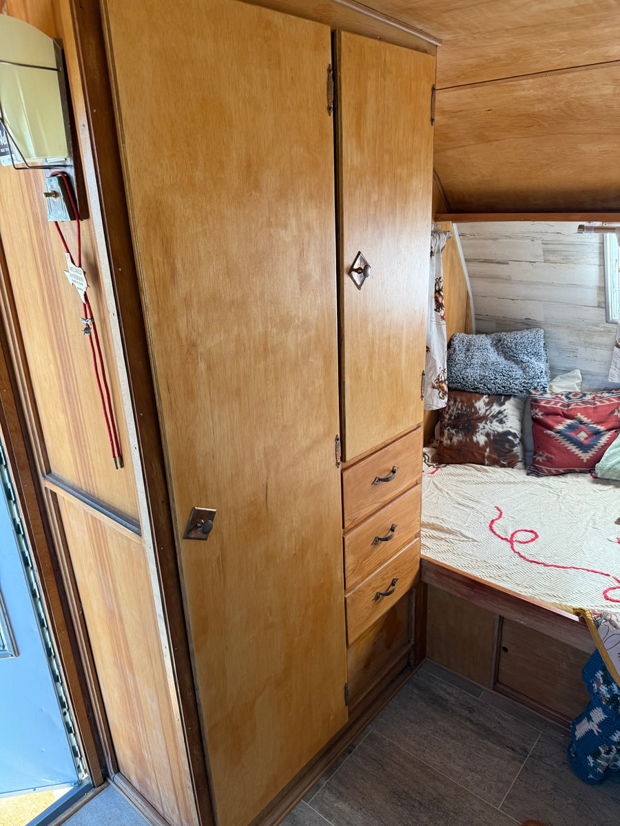 View of a vintage RV rental interior: features light wood cabinets, a cozy bed with decorative pillows, and tiled flooring.