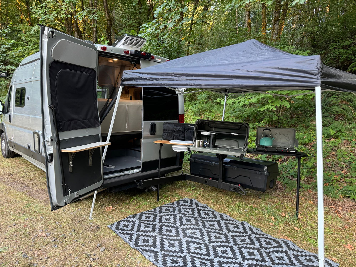 An RV rental camper van parked in a wooded campsite, featuring an outdoor kitchen, pop-up canopy, & ground mat.