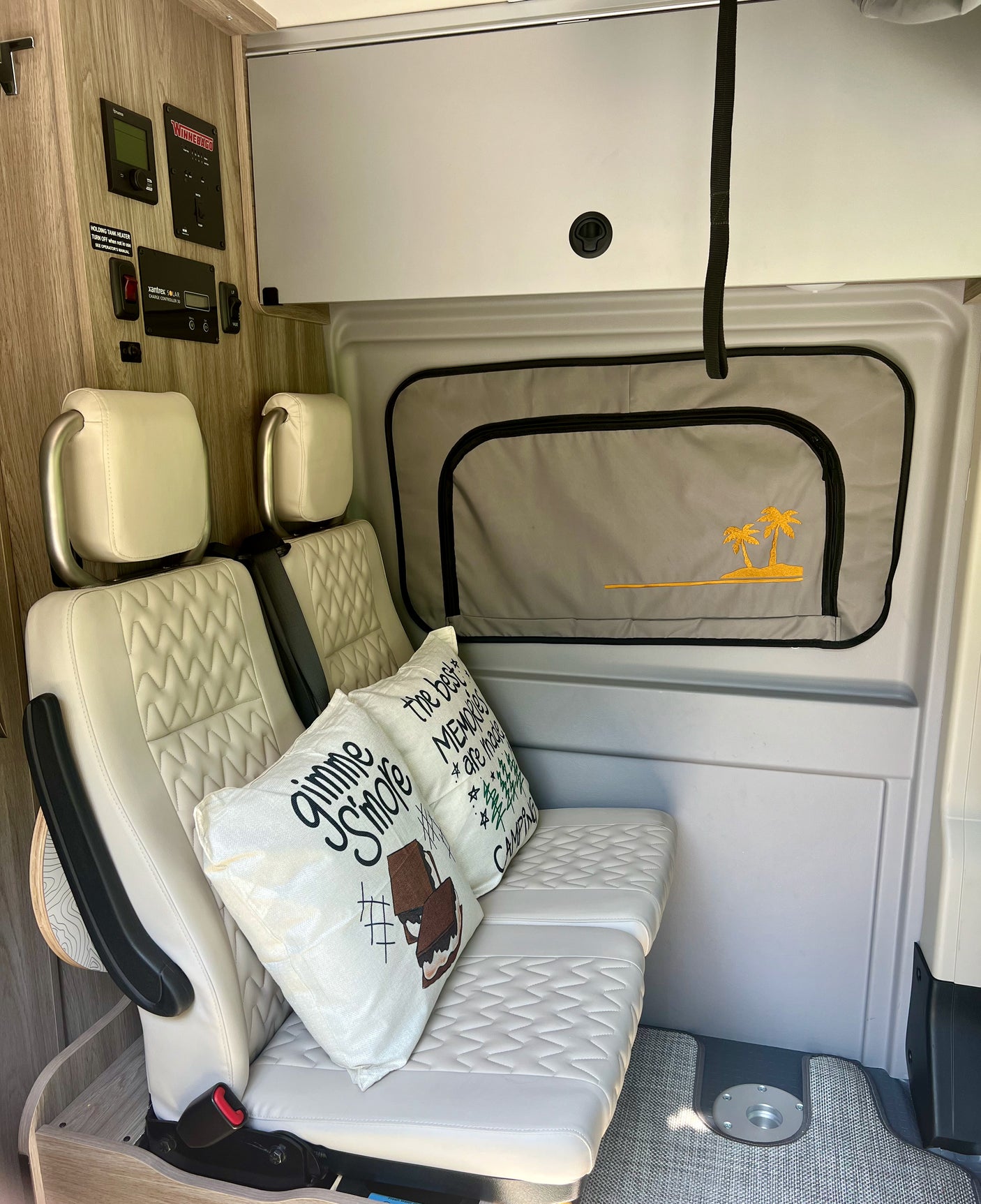 View of a modern RV rental interior featuring quilted seats, camping pillows, a control panel, and a palm tree window shade.