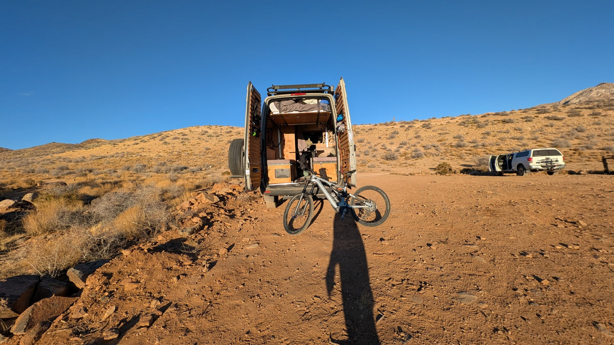 An RV rental camper van with open doors & mountain bike at camp. A person's shadow stretches across the desert ground with hills beyond. — photo 7