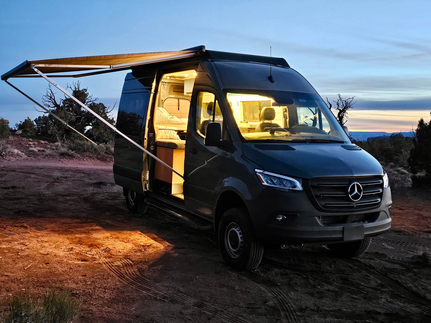 Cozy Mercedes-Benz Sprinter rv rental camper van with awning extended at sunset, showing illuminated interior.