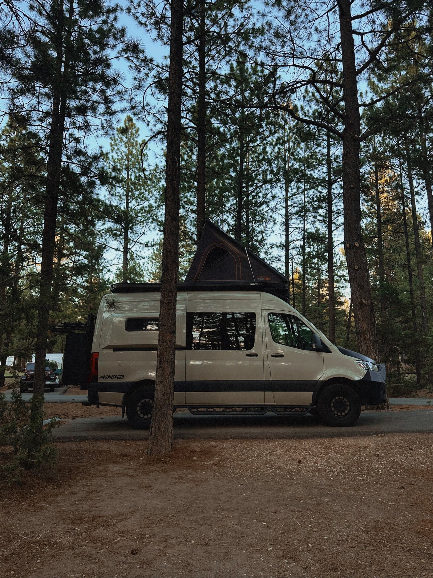 RV rental: A beige Mercedes-Benz Sprinter van with a rooftop tent, parked among tall pine trees at a serene campsite.
