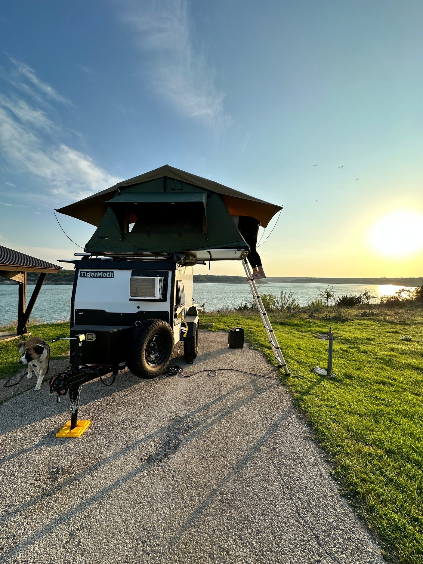 RV rental adventure: Lakeside sunset camping with a TigerMoth travel trailer, rooftop tent, and dog. Person on ladder.