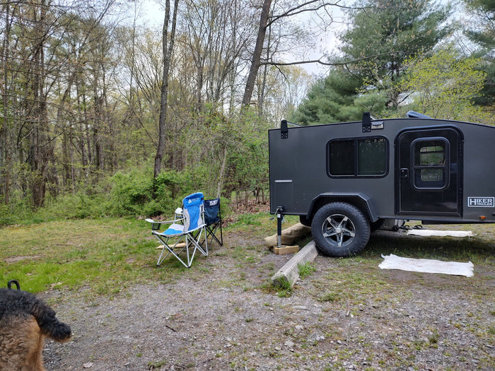 Hiker teardrop RV rental at a wooded campsite, chairs ready for relaxing. A dog's tail peeks in this outdoor scene.