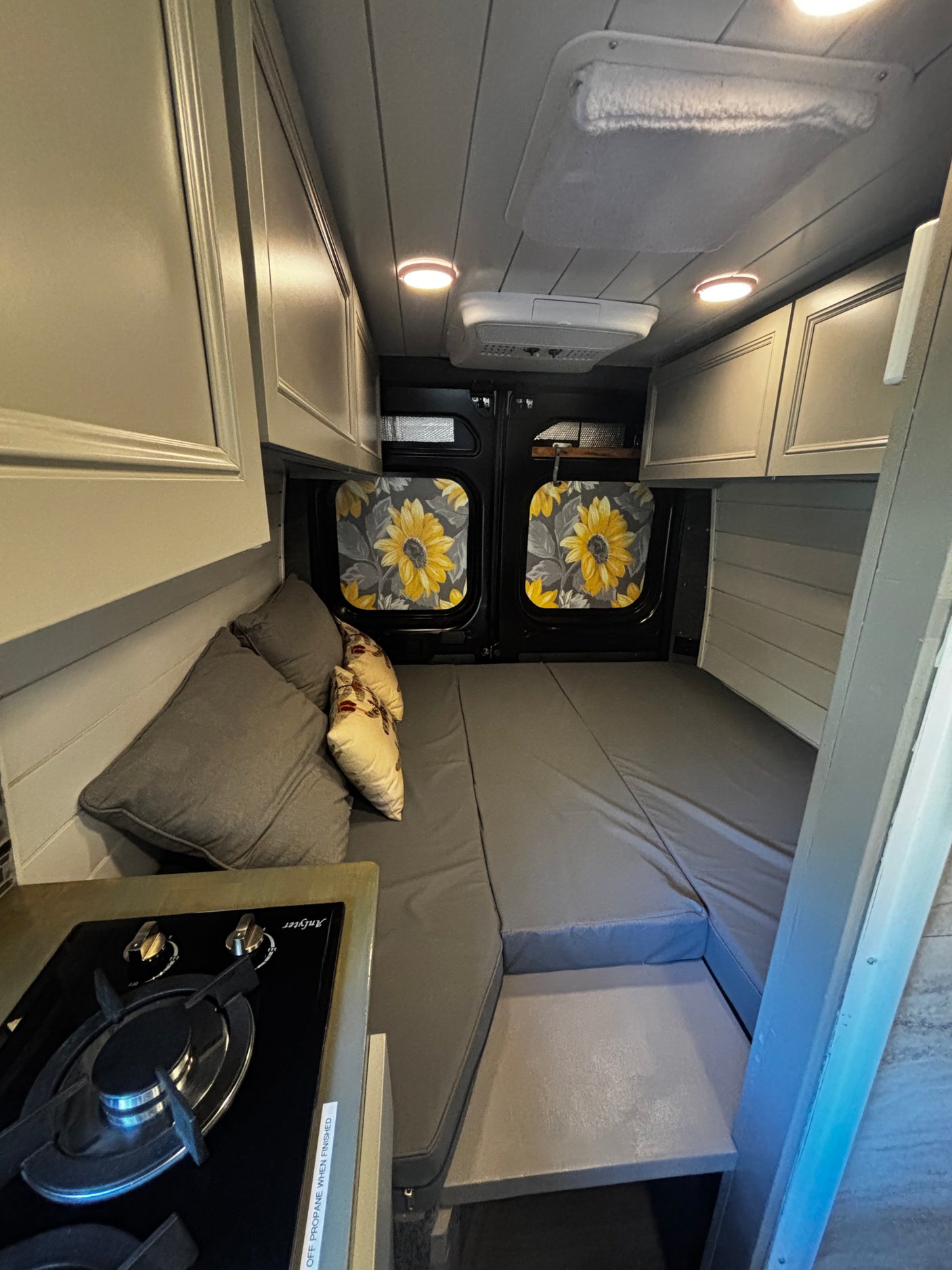 Cozy rv rental interior: kitchenette, bed with grey cushions & sunflower curtains, overhead cabinets, and ceiling lights.