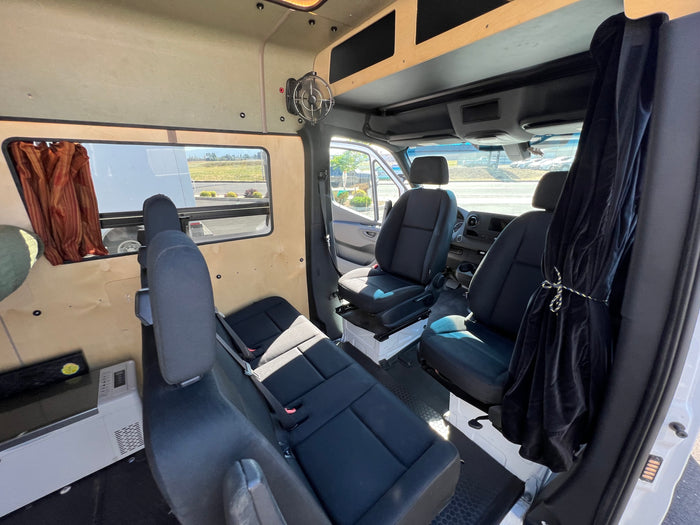 Interior view of an rv rental van, showing front and rear black cloth seating, light wood paneling, and a mounted fan.