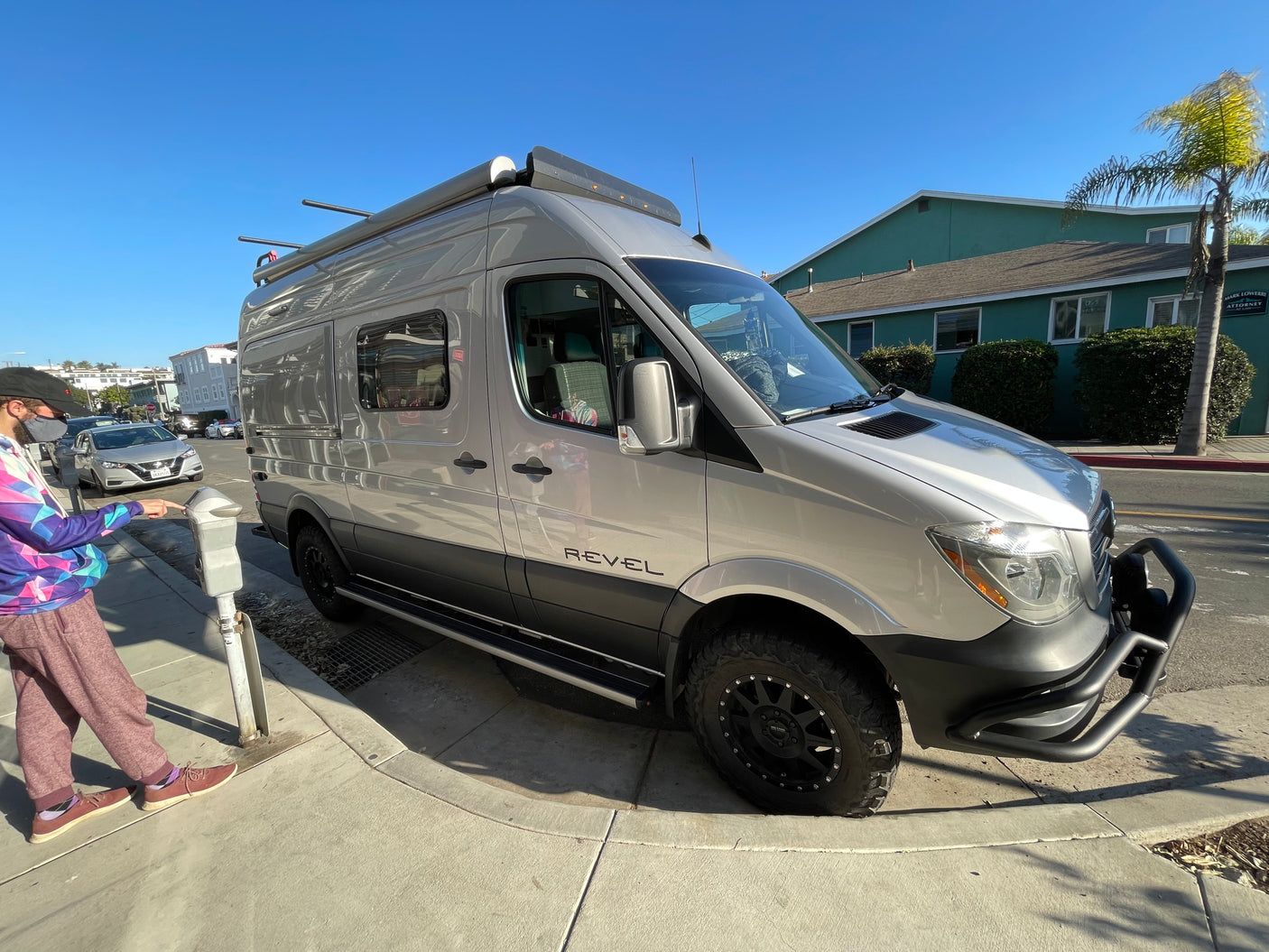 Shiny silver Winnebago Revel RV rental parked curbside on a sunny day, as a person operates a parking meter.