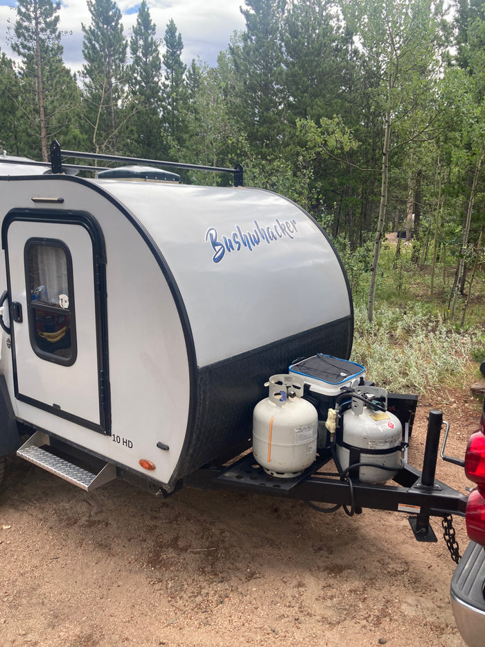Bushwhacker 10 HD travel trailer, an RV rental, with propane tanks at a wooded campsite.