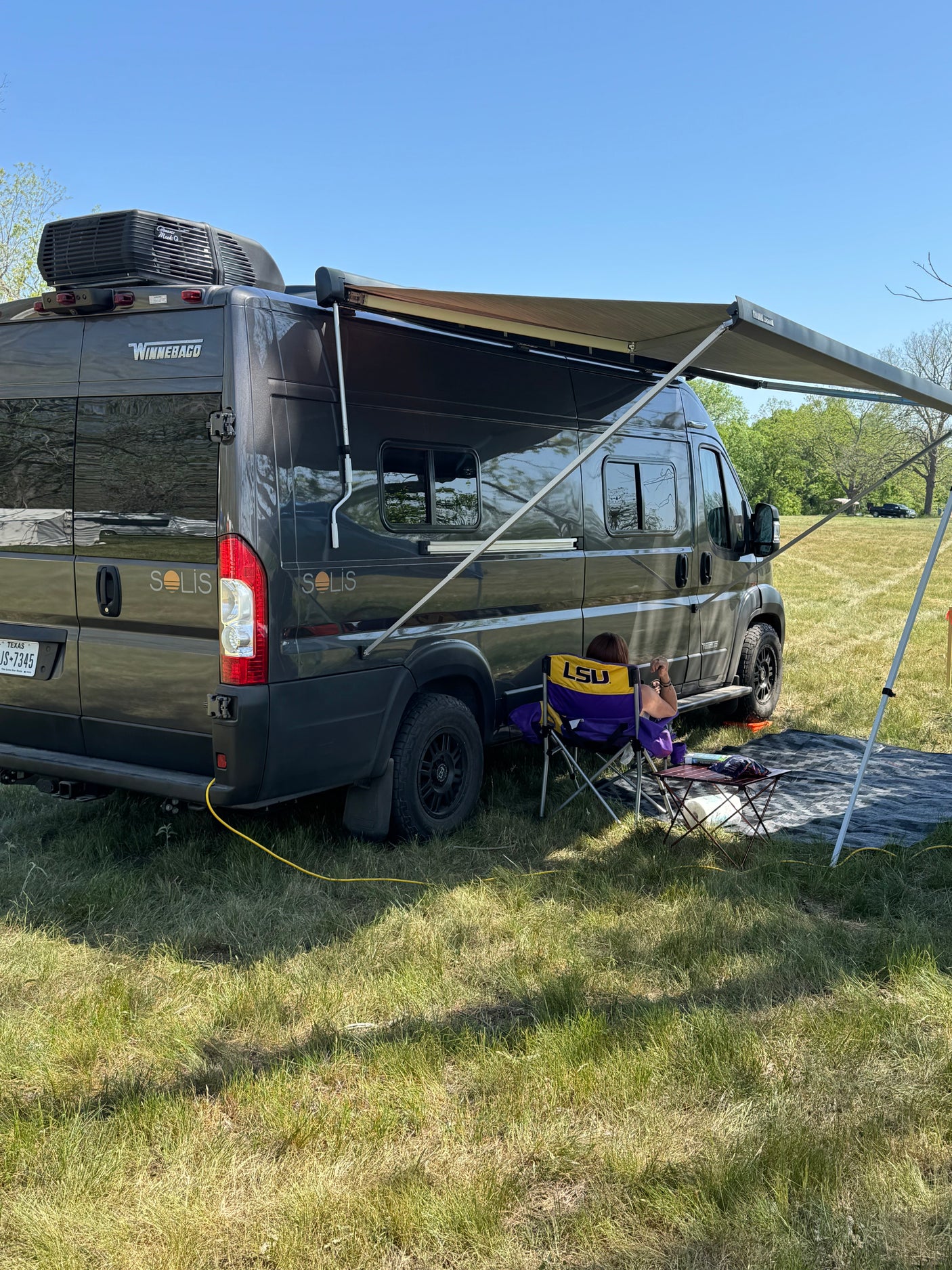 Relaxing RV rental setup: Winnebago Solis with awning deployed, person enjoying shade on LSU chair in a grassy field.