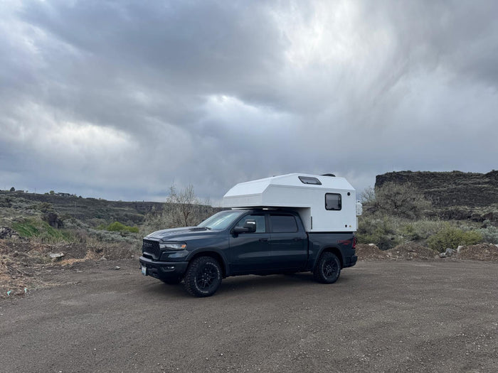 parked by tall pine trees and rocks under a blue sky.|Robust RV rental: a dark truck camper on a Ram pickup