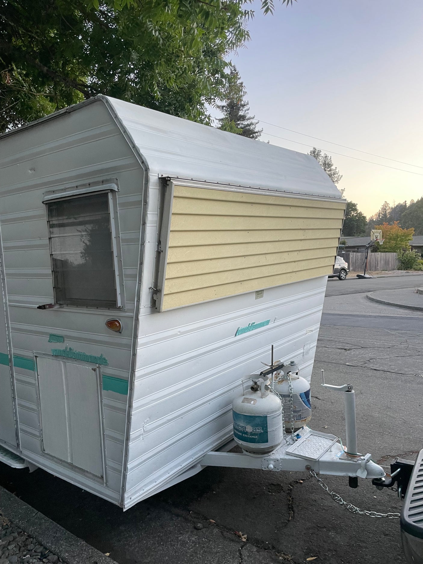 This vintage travel trailer could be your next RV rental! White camper with yellow side panel & propane tanks, parked outdoors.