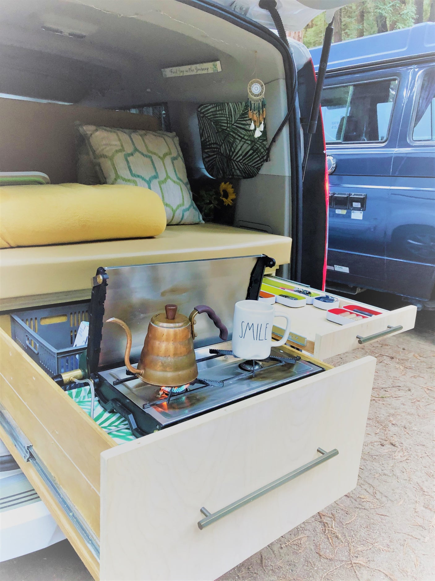 Inside a cozy RV rental: mobile kitchen with pull-out stove, kettle, 'SMILE' mug, & comfy sleeping area, perfect for camping.