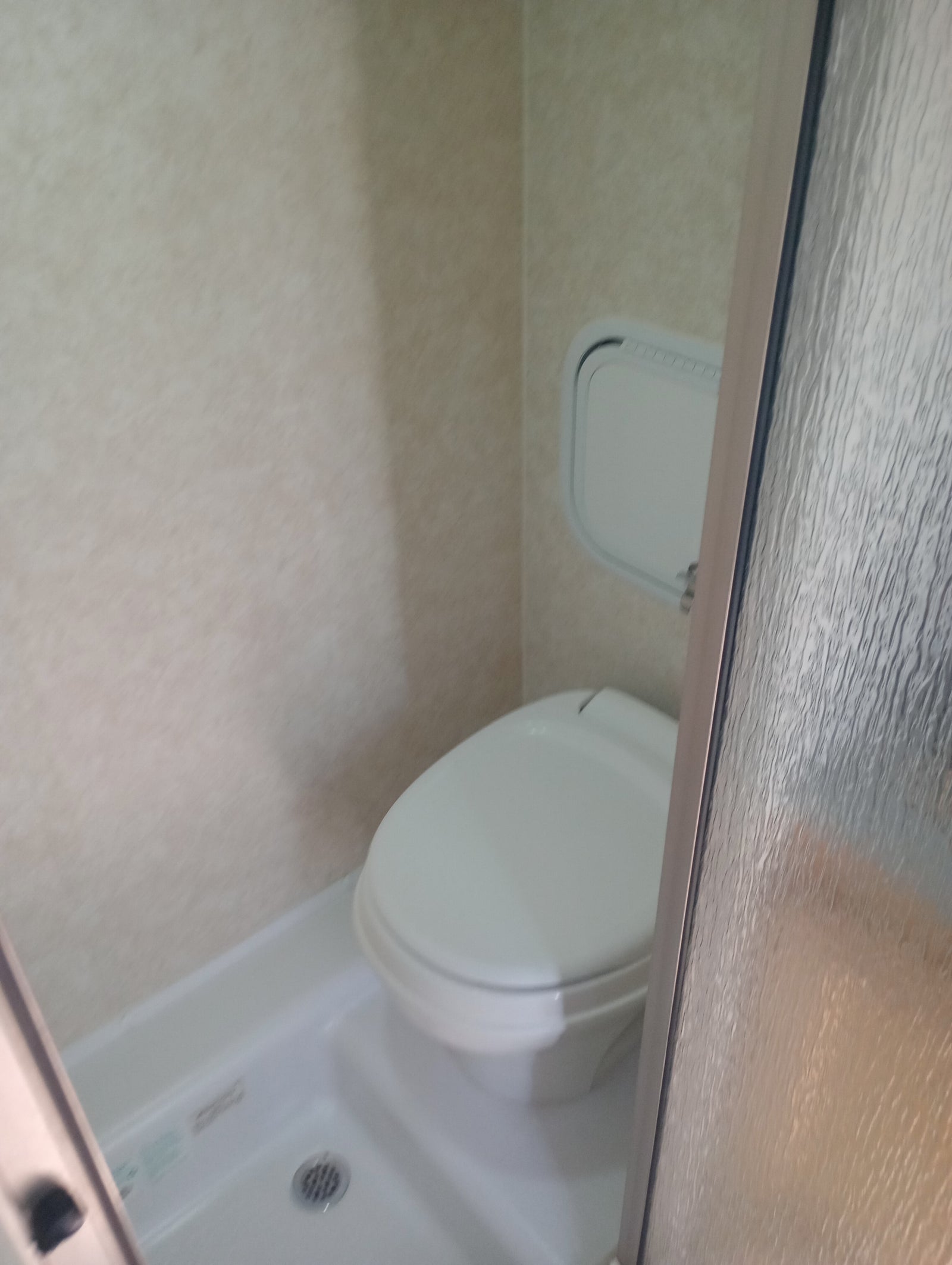 Compact RV rental bathroom featuring a toilet, shower, small wall panel, and textured glass door.