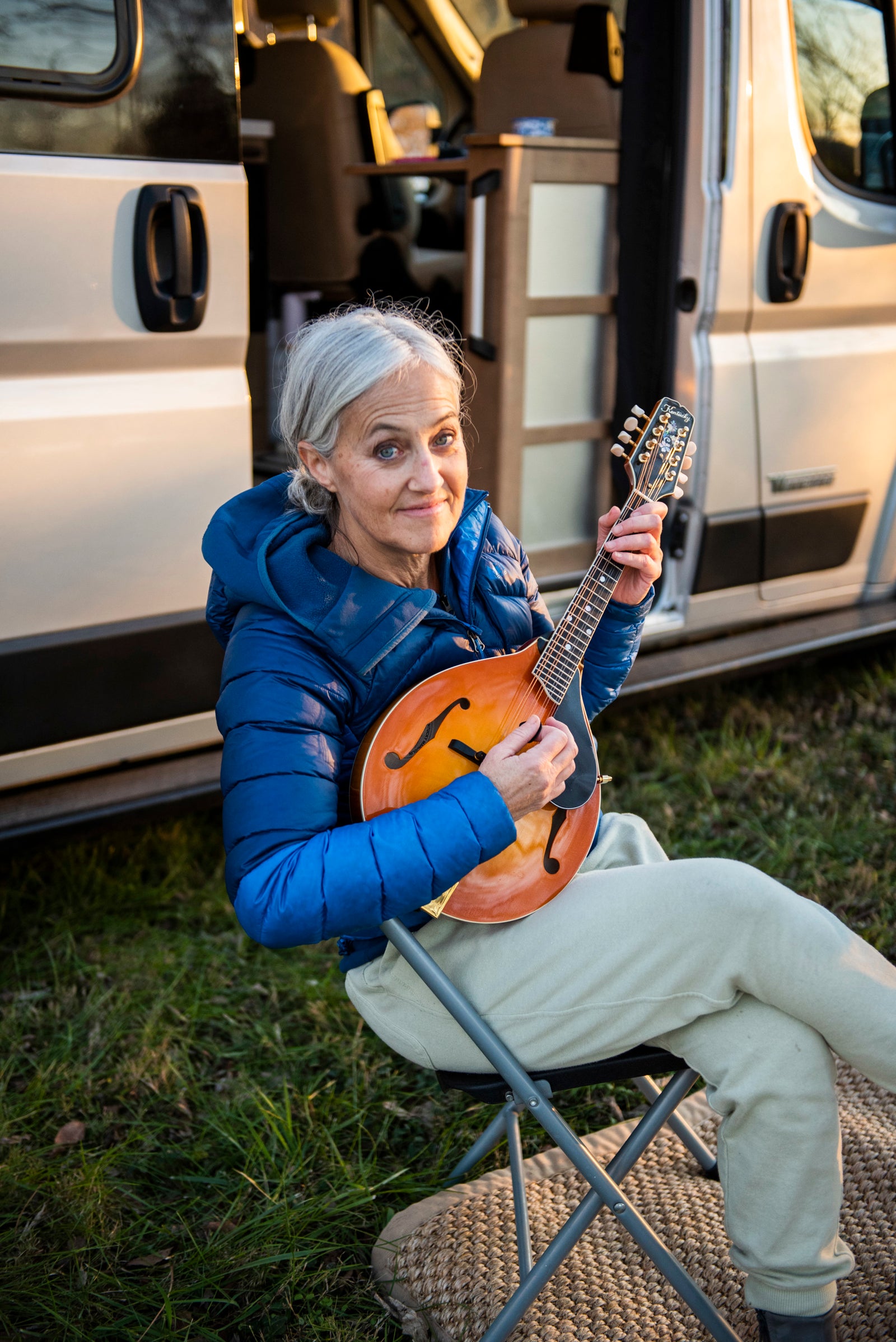 This smiling woman enjoys playing her mandolin outside her RV rental, sitting on a chair by the open camper van door.