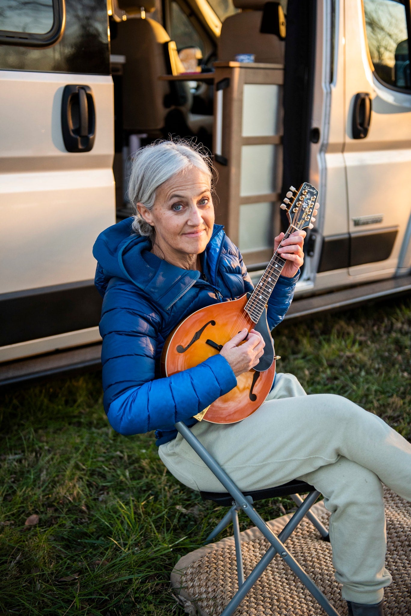 This smiling woman enjoys playing her mandolin outside her RV rental, sitting on a chair by the open camper van door.