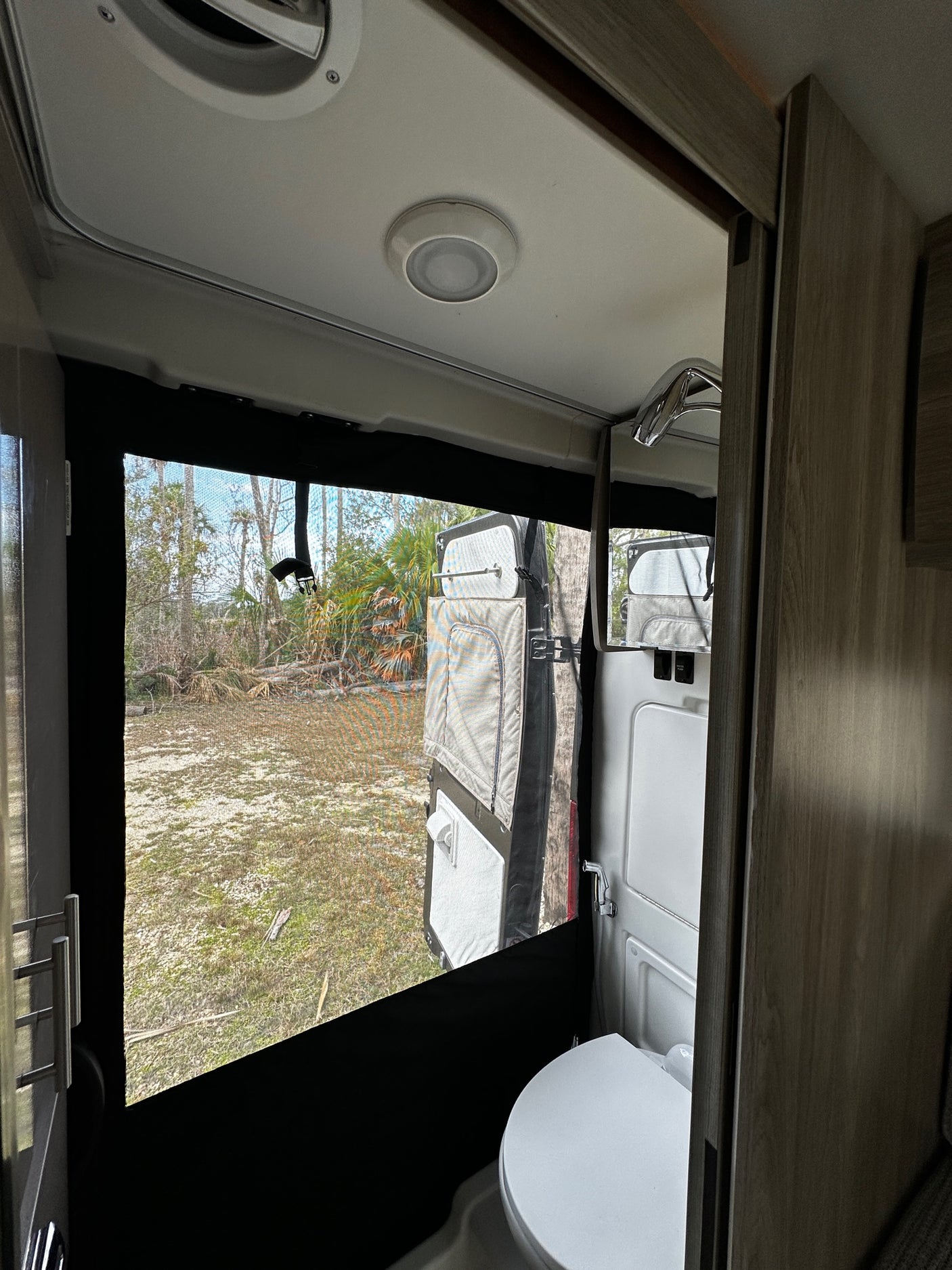 RV rental interior: Compact bathroom with toilet, shower faucet, and a window view of sandy terrain & trees.