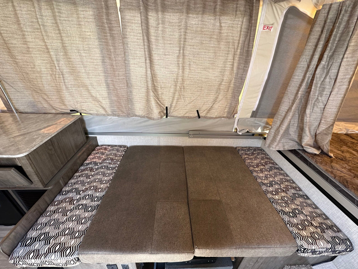 RV rental interior featuring a dinette with patterned cushions, transformable to a bed. Gray curtains and an exit sign visible. — photo 8