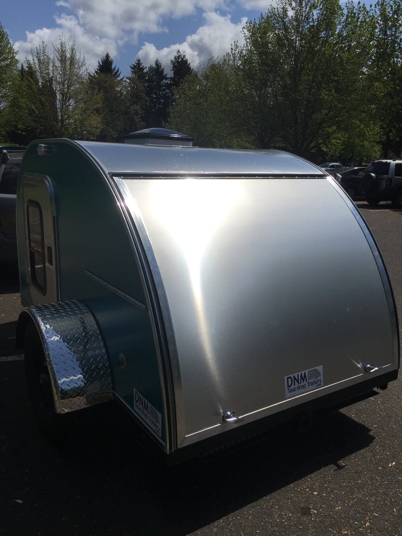 A stylish silver and teal teardrop trailer parked outdoors, reflecting sunlight. Consider this compact rv rental for your next trip.