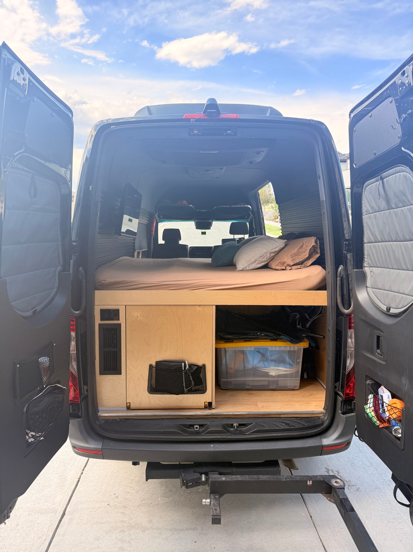 RV rental camper van interior. Features a comfortable bed, custom wood storage, and open rear doors, ideal for mobile living.