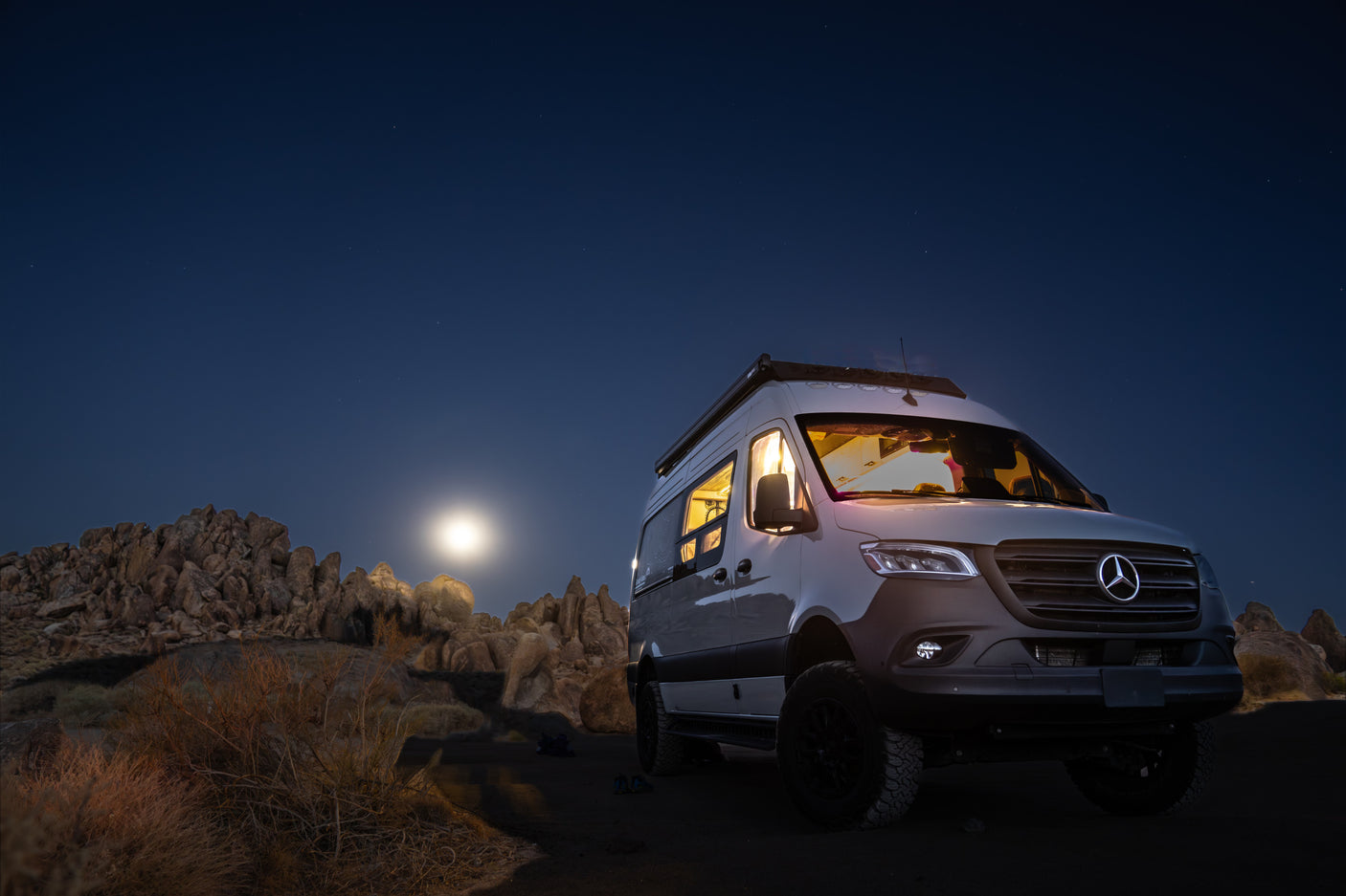 Moonlit desert camping with a bright Mercedes-Benz Sprinter RV rental under a starry sky. Interior lights glow invitingly.
