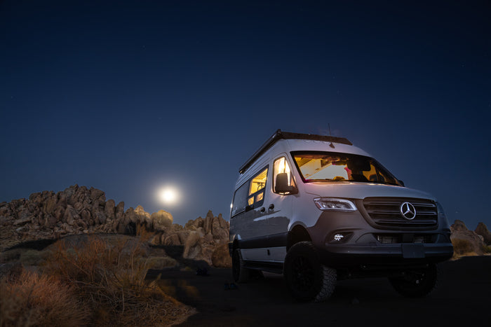 Moonlit desert camping with a bright Mercedes-Benz Sprinter RV rental under a starry sky. Interior lights glow invitingly.