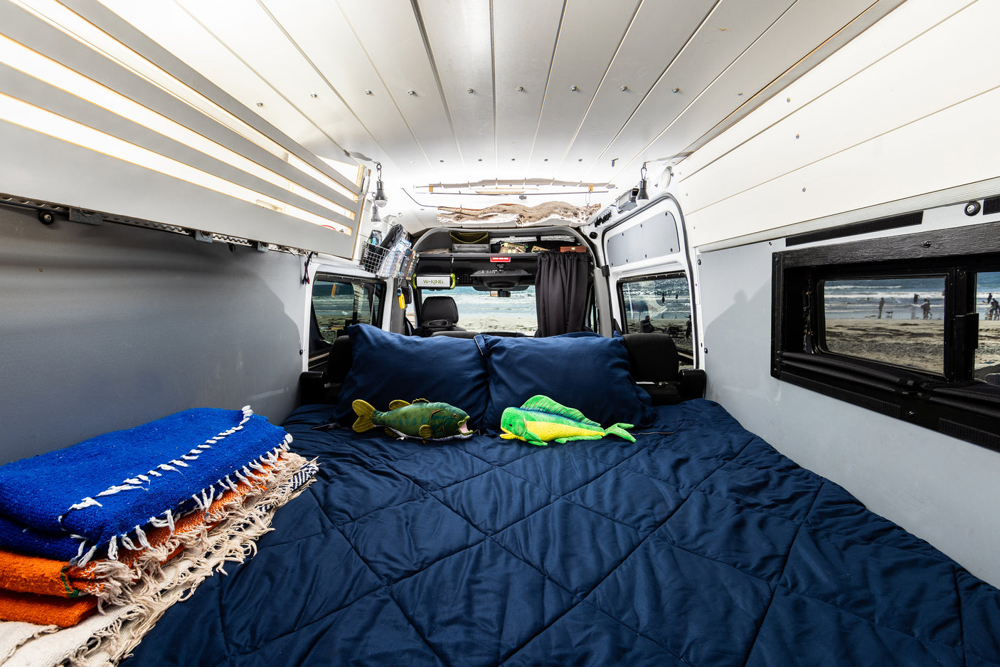 This cozy rv rental interior features a comfy bed with fish toys, stacked blankets, and open doors revealing a beach view.