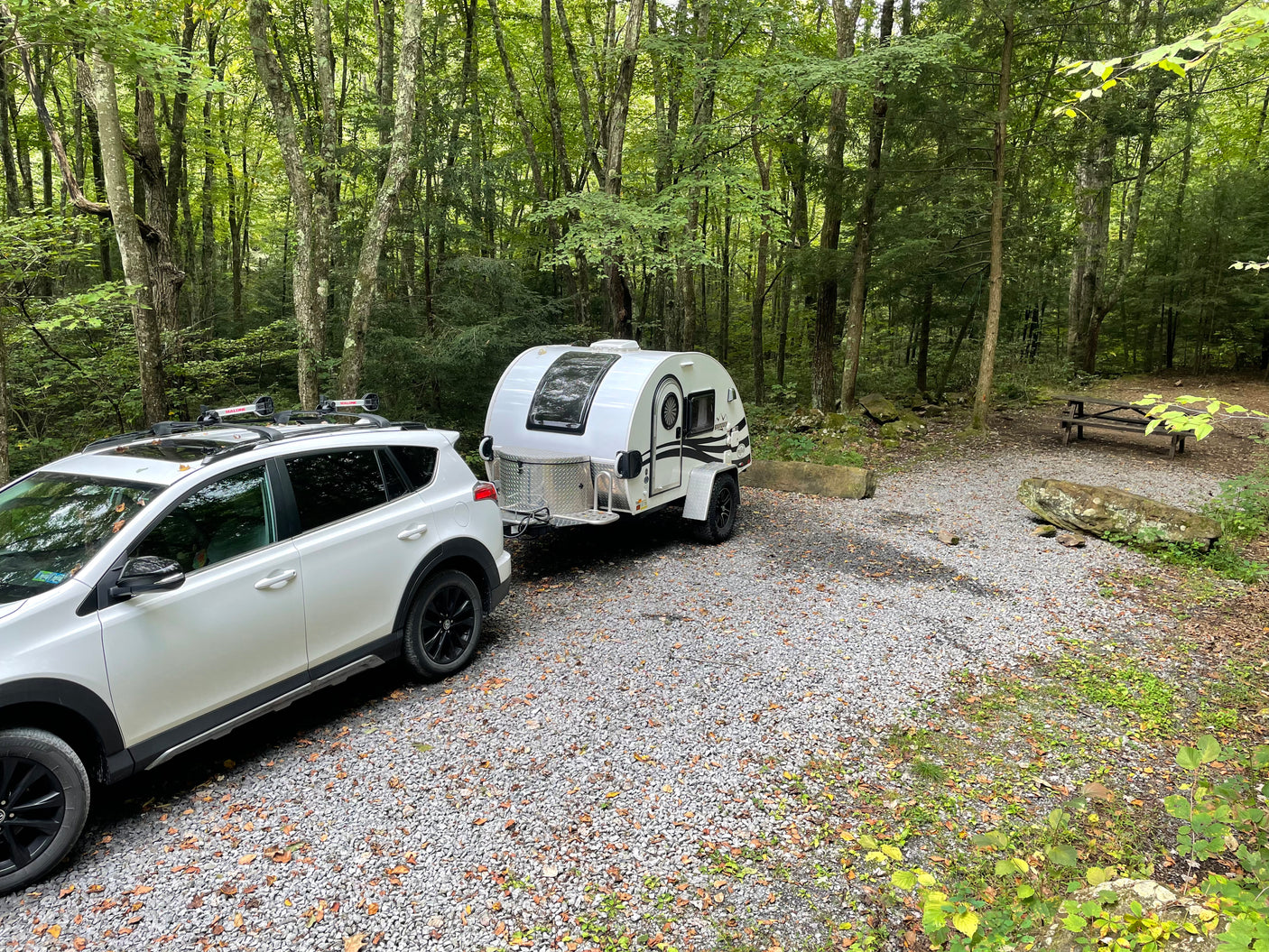 White SUV towing a teardrop RV rental at a wooded campsite with gravel, rocks, and picnic table.