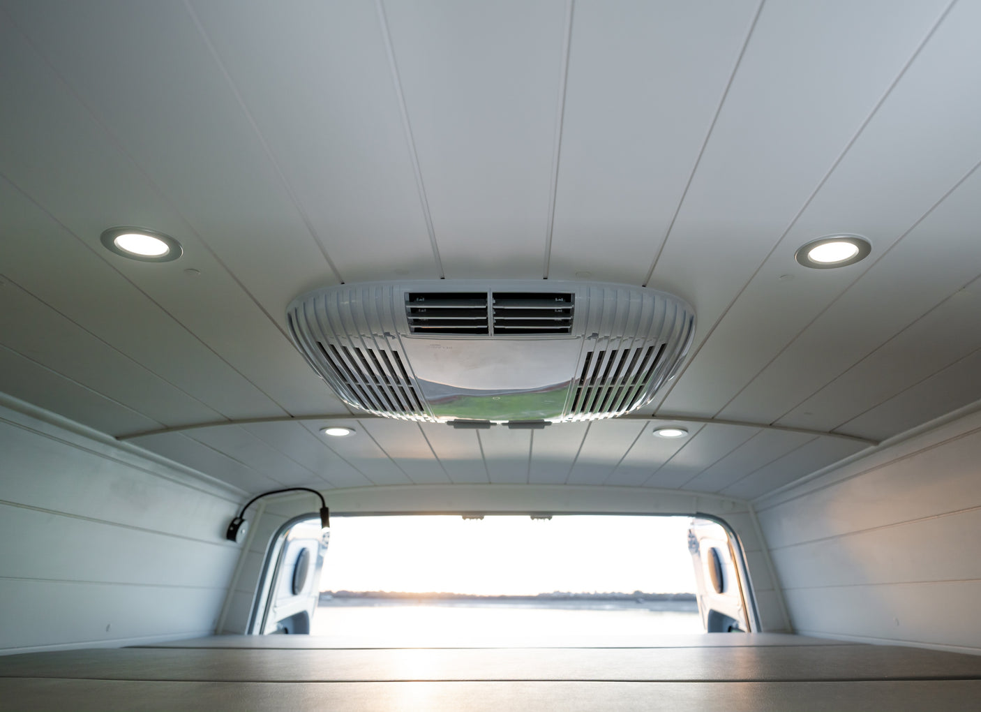 RV rental interior featuring a white paneled ceiling with AC unit, recessed lights, and a cozy bed looking out bright open rear doors.