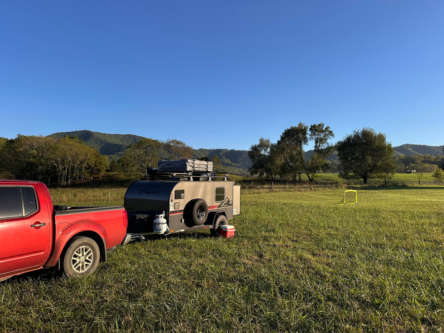 Red pickup truck and gray **RV rental** camper trailer set up in a vast green field with mountains & clear skies.|Electric truck tows an off-road camping trailer through a sunny forest. Great for an RV rental adventure!|Cozy RV rental interior: A bed with orange bedding