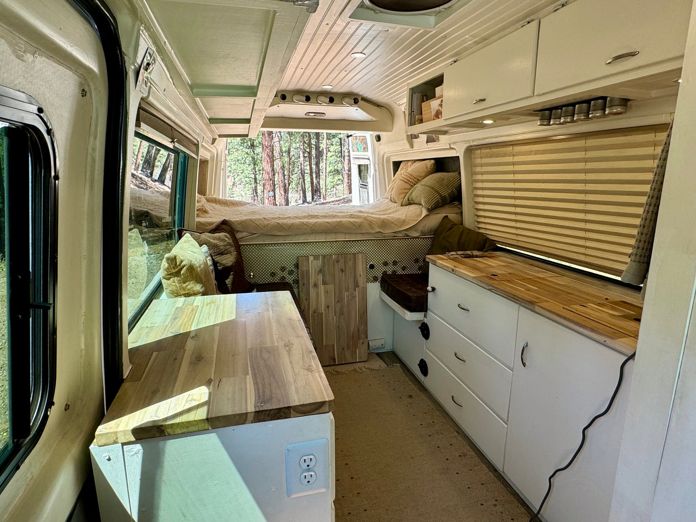 Cozy RV rental interior: bed with pillows, wooden countertops, kitchen cabinets, and a scenic forest view.