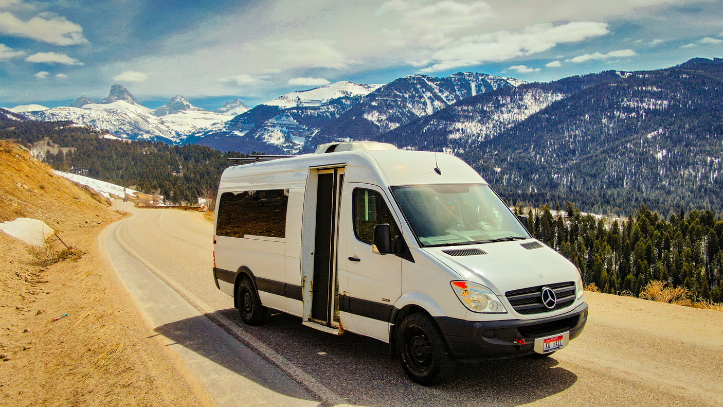 White Mercedes Sprinter RV rental on a scenic mountain road, featuring snow-capped peaks and pine forests.