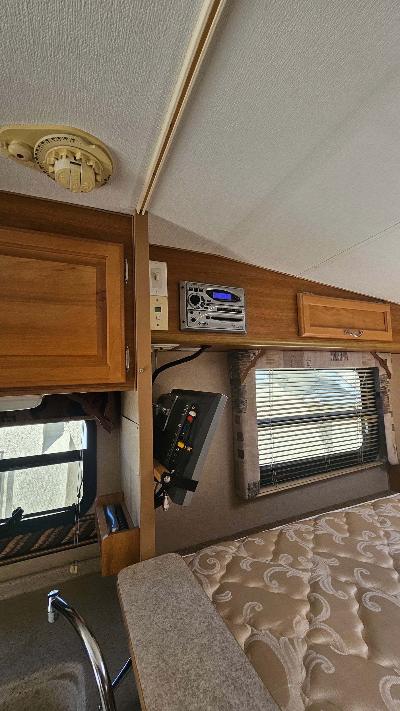 Inside a compact rv rental: wood cabinetry, Jensen entertainment system, a window, patterned bed, and kitchen area.