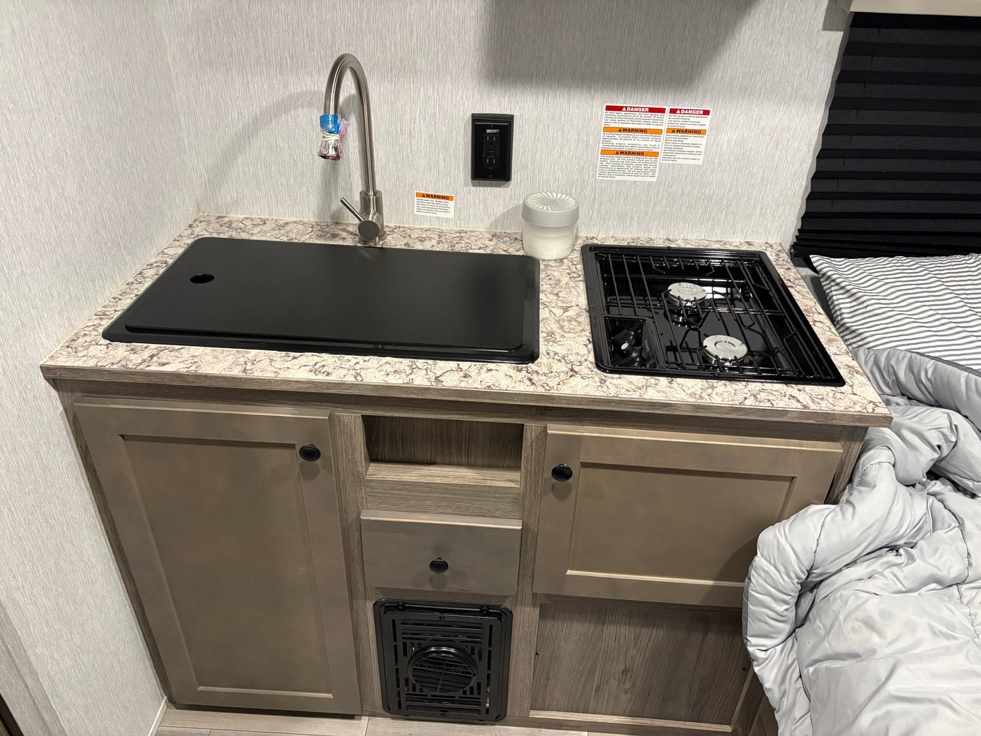 Compact RV rental kitchen with a covered sink, faucet, two-burner stove, and lower cabinets. A bed with comforter is on the right.