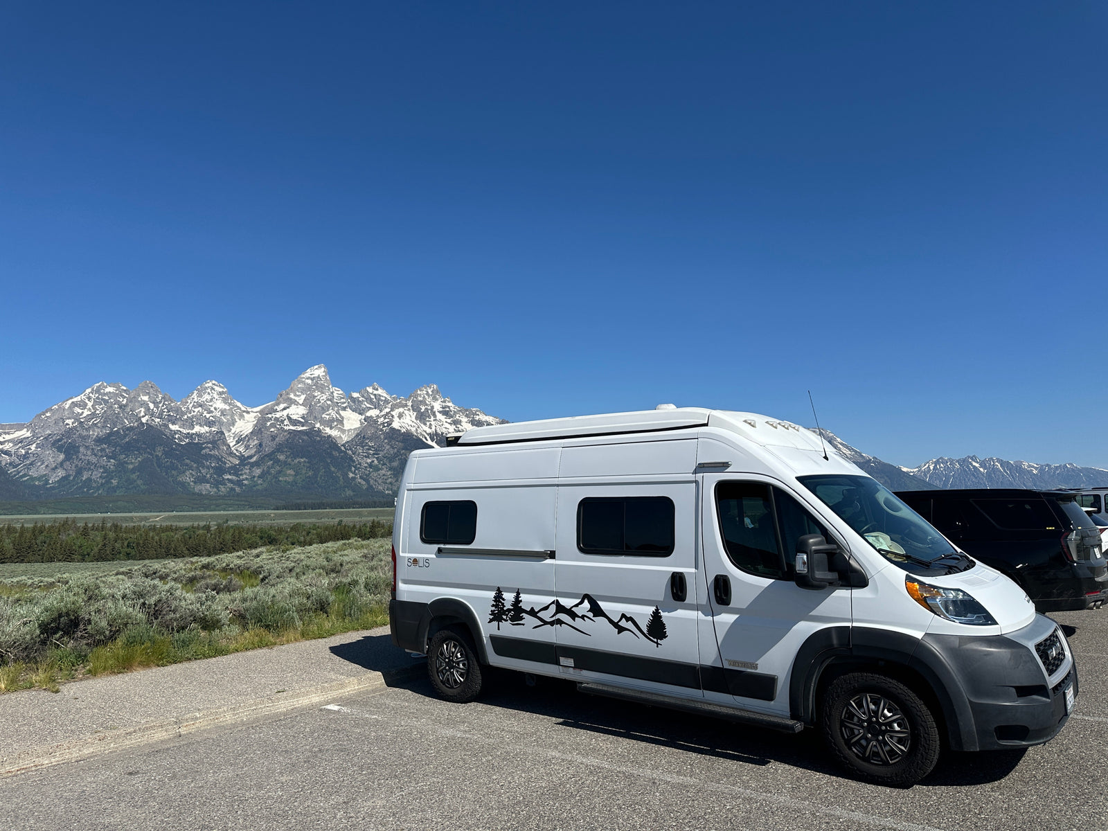 Scenic RV rental adventure! White camper van parked with stunning snow-capped mountains in the background under a clear blue sky.