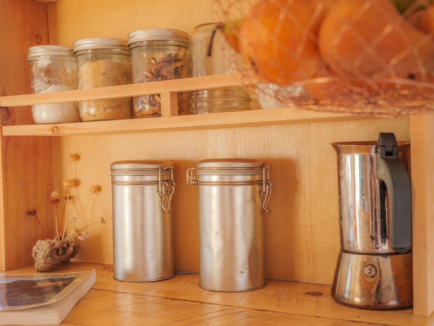 Cozy RV rental kitchen: wooden shelves hold jars of ingredients, metal canisters, a moka pot, and fruit in a basket.