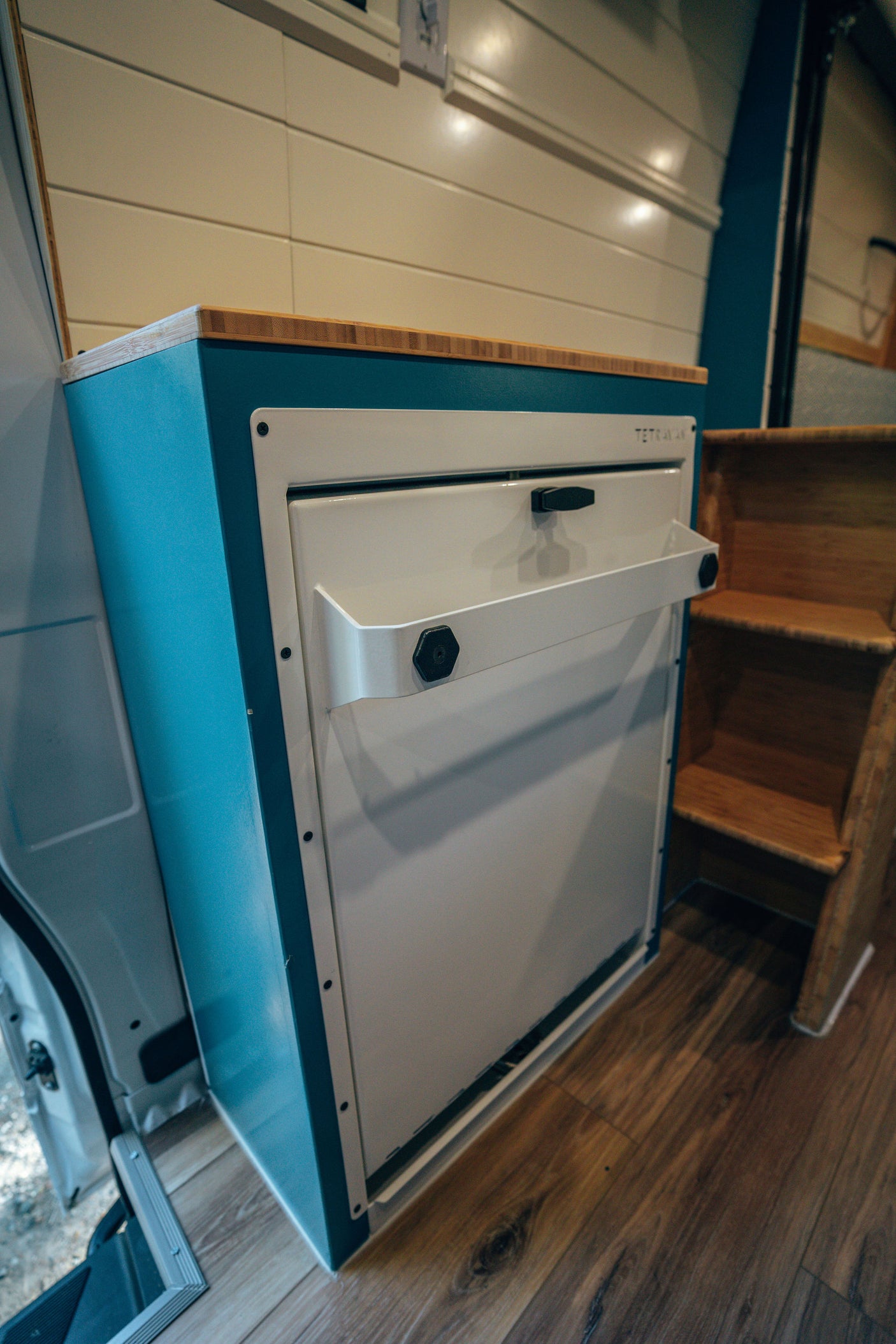 RV rental interior: TETRA VAN turquoise cabinet with wood top, likely a fridge, and laminate floor in a cozy campervan.
