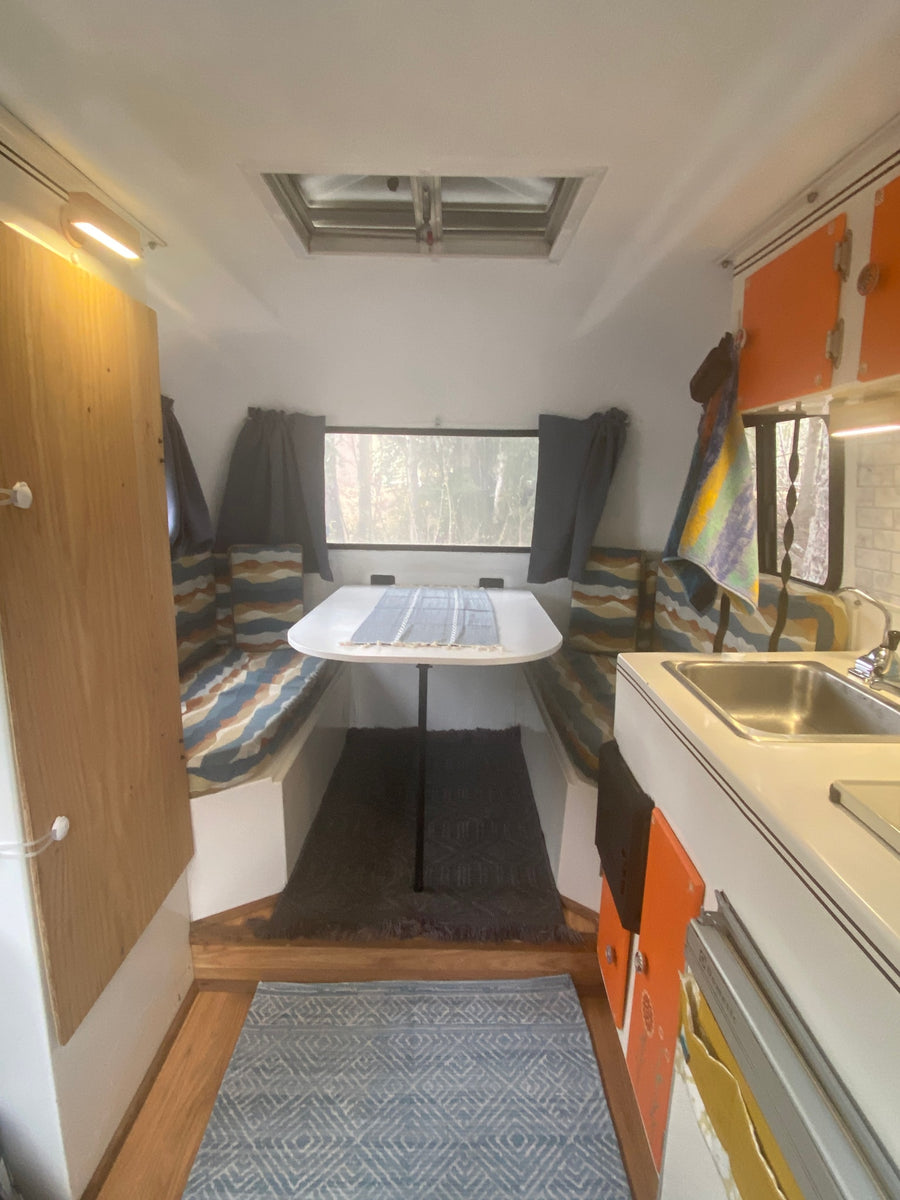 This cozy RV rental interior features a dinette with patterned cushions, a compact kitchen, and a window revealing trees.