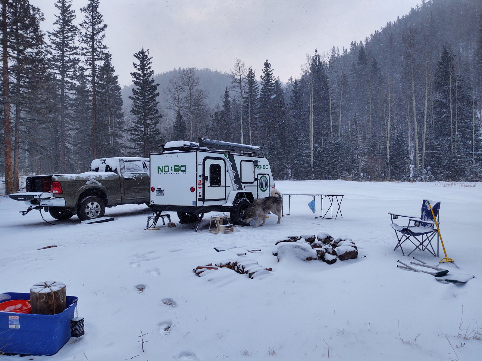 Winter RV rental adventure: A truck and NOBO camper trailer parked in a snowy forest, with a dog, fire pit, and gear.