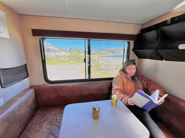 Relaxed woman reading in an RV rental dinette overlooking a scenic beach. Perfect for your next travel adventure.