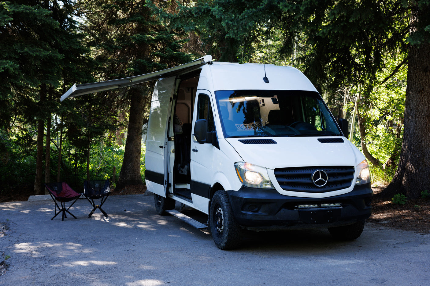 Enjoy nature with this Mercedes Sprinter rv rental camper van. Awning extended, chairs out, nestled among green trees.