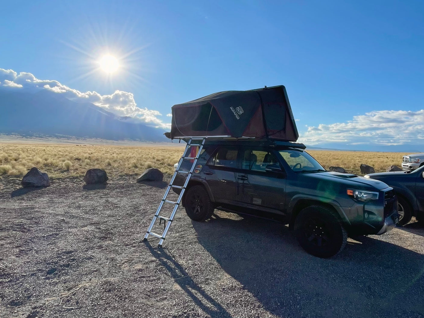 An RV rental SUV with a rooftop tent & ladder set up for camping, with sunny mountains & golden fields in the background.
