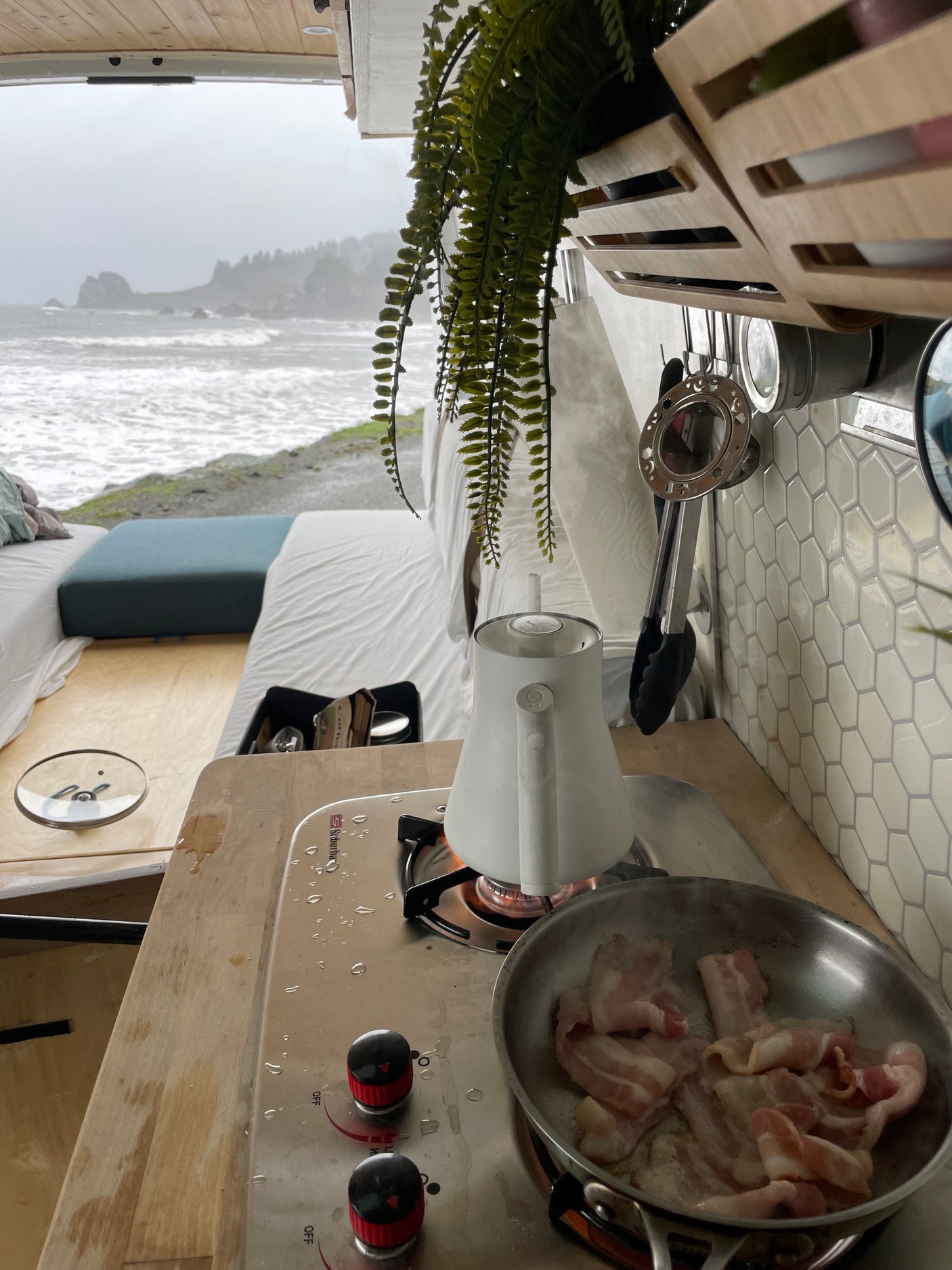 Inside an **rv rental** van, breakfast bacon cooks on the stove beside a boiling kettle, offering a cozy view of the stormy ocean.