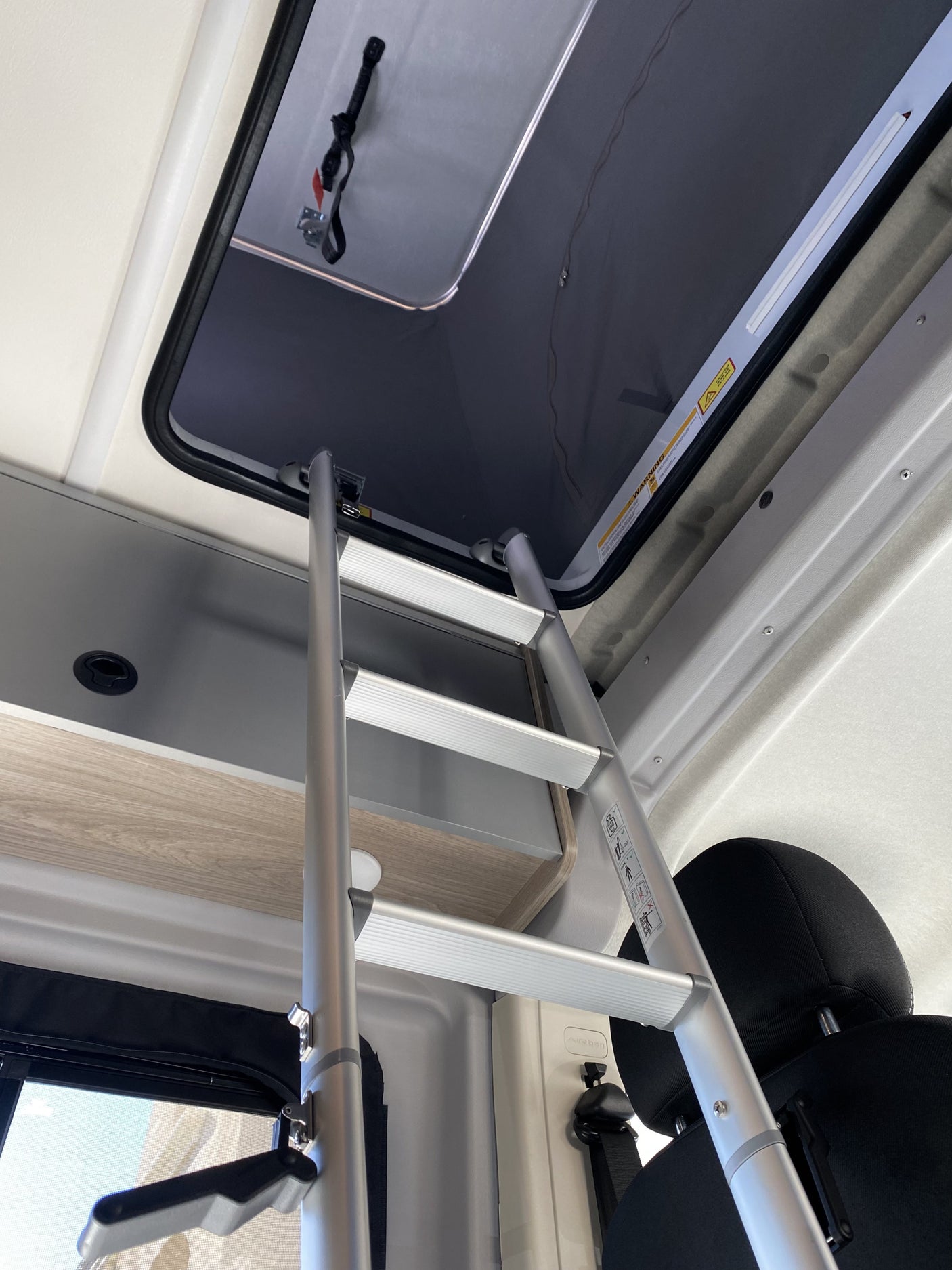 Interior of an RV rental: A silver ladder provides access to an upper sleeping bunk, with a roof hatch and storage units.