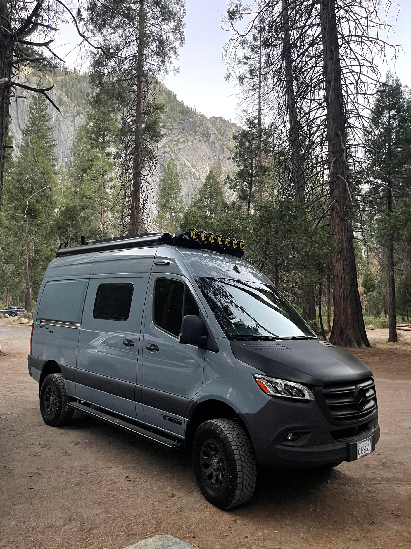 Adventure-ready gray Winnebago Revel RV rental in a mountain forest. Tall pines frame the customized van with roof lights & off-road tires.
