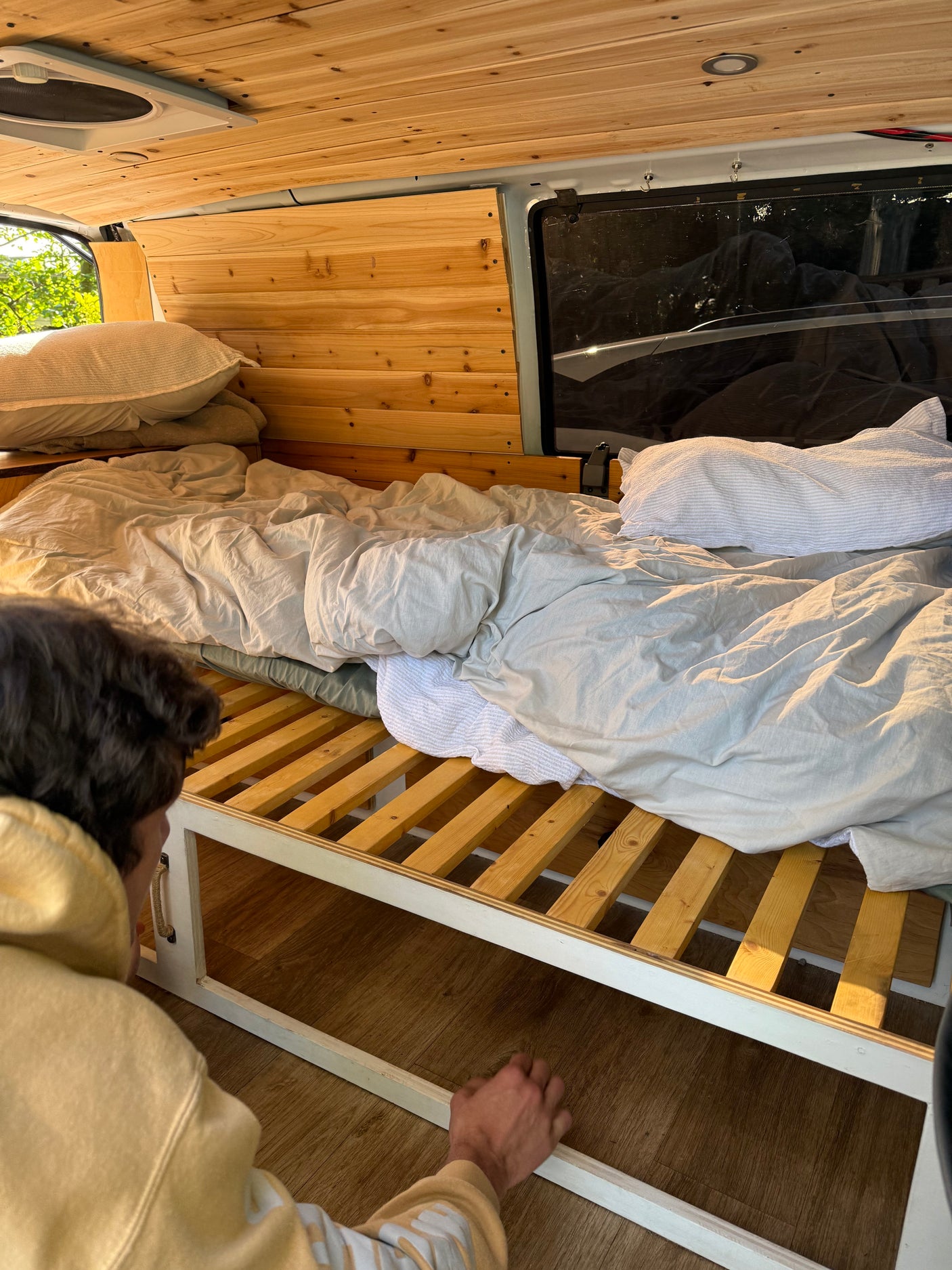 Cozy RV rental bed with wooden slatted frame and a person accessing under-bed storage in a camper van.