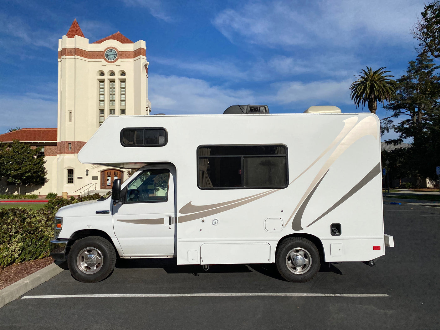 White Class C RV rental parked for travel next to a grand building with a clock tower.