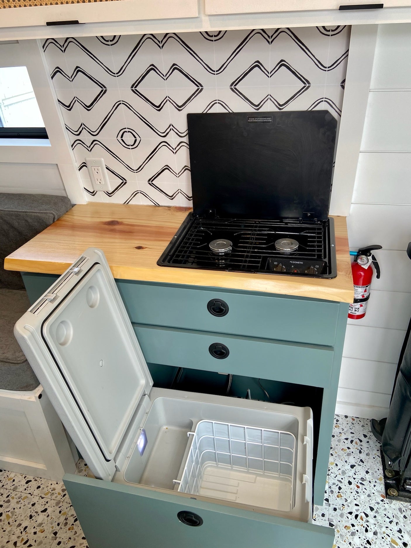 Compact RV rental kitchen featuring a two-burner stove, pull-out fridge, and fire extinguisher, set against stylish backsplash.