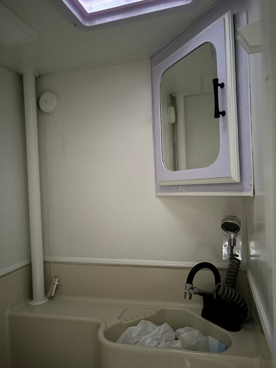 Compact RV rental bathroom: sink with pull-out black faucet, wall-mounted shower, mirrored cabinet, and skylight. Bags in sink.