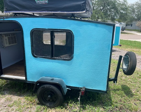 Light blue teardrop RV rental: compact camper with a roof-top tent, AC unit, and rear spare tire. Adventure-ready.