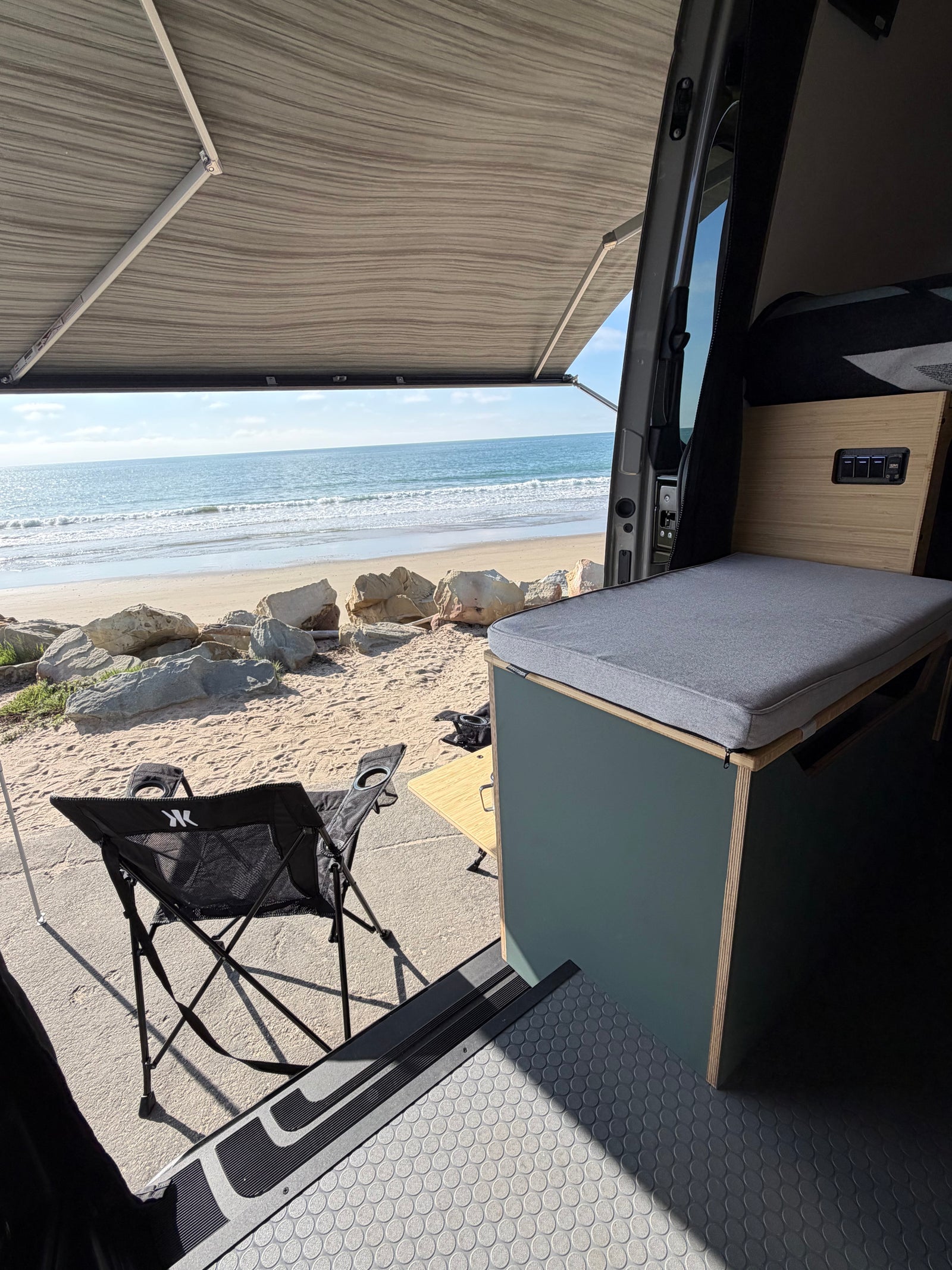 Beachfront RV rental: open camper van with awning, indoor seating, and outdoor chair overlooking the ocean and sandy beach.